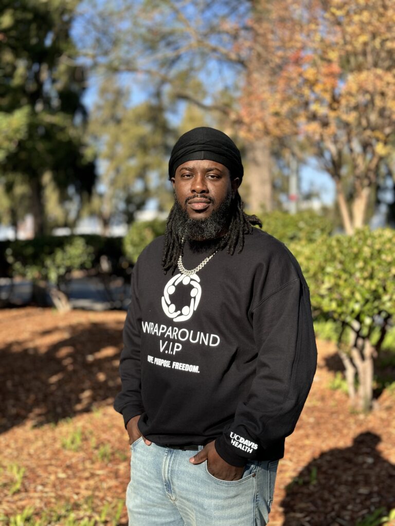 UC Davis Health Wraparound Violence Prevention Professional and TSN Coordinator, Chevist Johnson standing and posing on a sunny day with slightly blurred trees, bushes and woodchipped lawn in background.